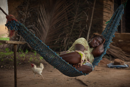 Yongoro, Sierra Leone - June 03, 2013: West Africa, the village of Yongoro in front of Freetown, young woman resting on a hammock in the background passes a chickenのeditorial素材