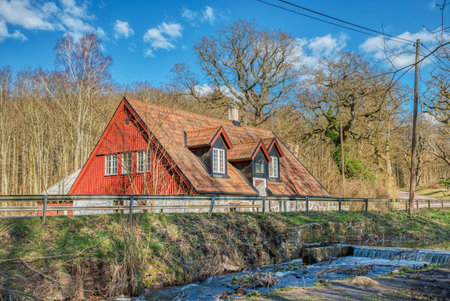 Detached rural red countryside house with pitched roof by a river with a short waterfall. Farmer house with orange roof by a countryside road in the woods. Countryside and isolated lifestyle conceptの写真素材