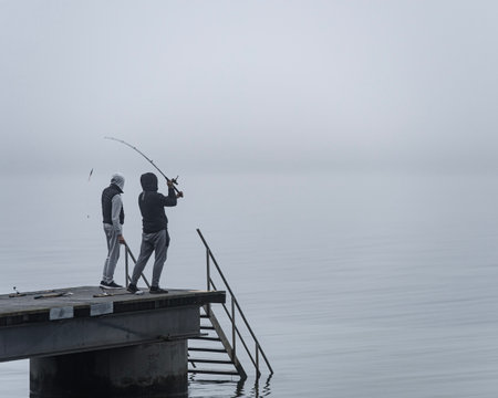 2 unrecognisable people from behind fishing in the fog on a pier in Klagshamnsstranden or Klagsham strand. An unrecognisable guy is throwing the bait with a fishing rod in the mist on a gloomy dayの写真素材