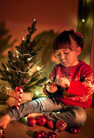 Holiday preparation and festive decor. A girl in a red sweater sits by a small, decorated Christmas tree, holding an ornament. Child in a cozy setting exudes a joyful and magicalの写真素材