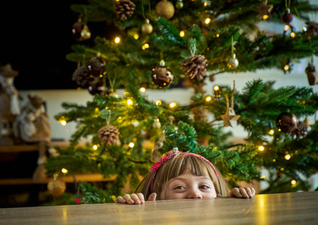 A curious child peeking over a festive table, with a Christmas tree in the background, captures the magic, warmth, and joy of the holiday season. A girl swung with anticipationの写真素材