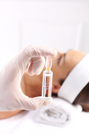 Gloved hand medical shows ampoule with a cosmetic preparation, in the background a woman waiting for surgeryの写真素材