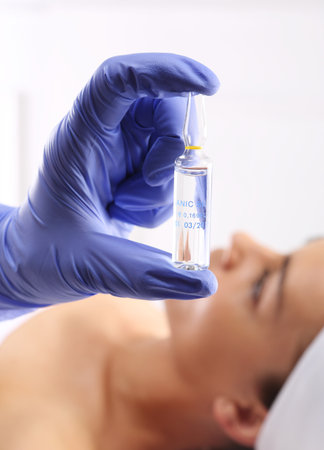 Gloved hand medical shows ampoule with a cosmetic preparation, in the background a woman waiting for surgeryの写真素材