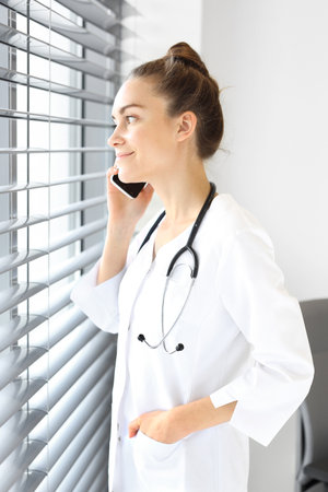 The doctor dressed in a white lab coat talking on the phone while standing at the window.の写真素材
