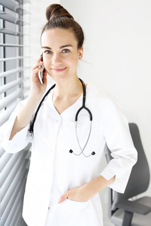 Attractive physician talking on the phone. The doctor dressed in a white lab coat talking on the phone while standing at the window.の写真素材