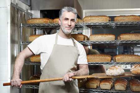 Baker in the bakery. A handsome baker with a loaf of fresh bread.の写真素材