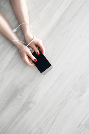 Dependence to a mobile phone. Hands of a woman connected by a cable charger are holding a mobile phone.の写真素材