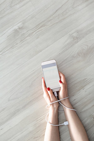 Addiction to the phone. Hands of a woman connected by a cable charger are holding a mobile phone.