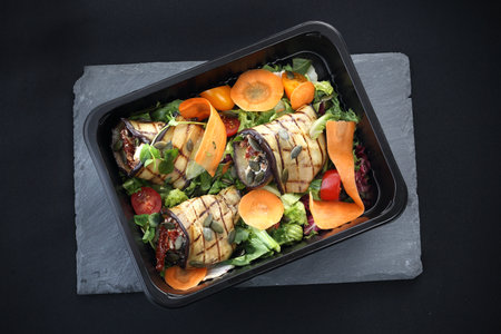 Grilled aubergine rolls with dried tomatoes and cheese served on lettuce and carrots packed in a container for food.の写真素材