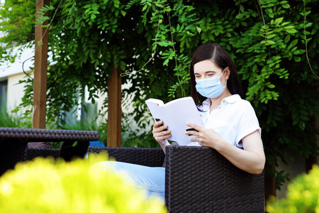 A woman in a mask is reading a book while sitting in a cafe. A lonely woman reads a book maintaining a social distance.の写真素材