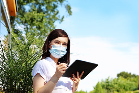 Remote work during an epidemic. A woman freelancer in a protective mask works in an urban space using a tablet.の写真素材