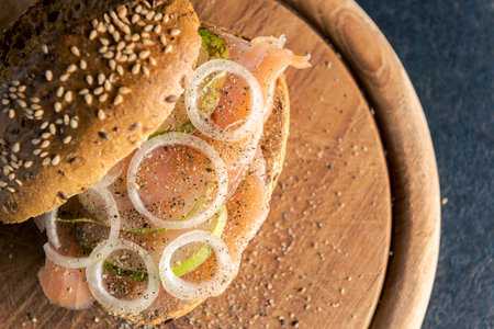 Top view of whole-grain bun with wild salmon, onion rings and slices of pear on wooden board and...