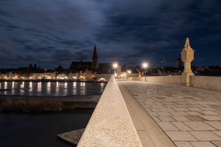 Historic illuminated stone bridge in Regensburg, Bavaria with cathedral in background at nightの写真素材