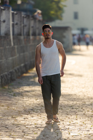 Full-length shot of young arabic man in white sleeveless shirt with sunglasses walking down a street...