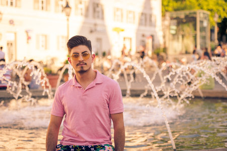 Urban portrait of young arabic man in colorful summer outfit with sunglasses sitting next to waterspout fountain on crowded public square on hot summer day smiling at cameraの写真素材