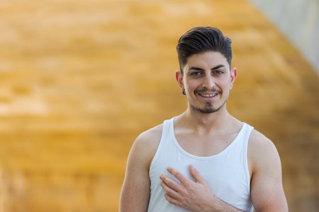 Young smiling arabic man in summer outfit with sleeveless shirt standing under arch of sunlit stone...