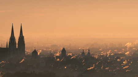 Overview of city skyline at the beginning of the heating period in autumn with many smoking chimneys in morning backlight twilight in Regensburg, Bavaria, Germanyの写真素材