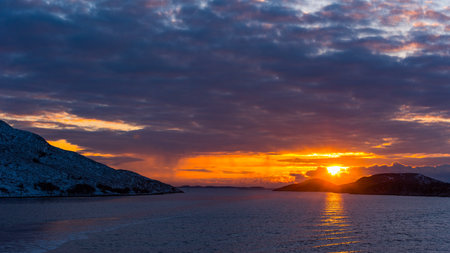 Glorious sunset over norwegian coast with snow covered rocks and dense clouds seen from open seaの写真素材