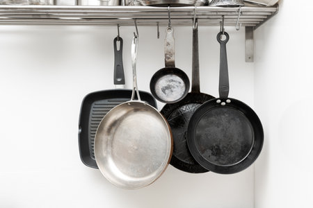 Various pans in different sizes and forms for cooking and frying hanging on metal hooks from shelf in kitchen with white wall in backgroundの写真素材