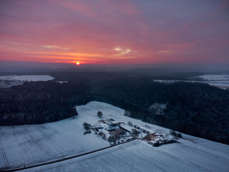 Drone panorama of amazing red sunrise behind clouds over bavarian forest and isolated farm area with snow in winterの写真素材