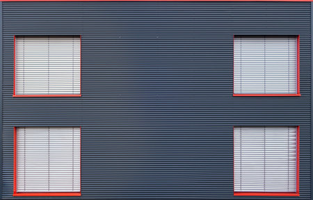 Four, with gray blinds closed windows with red frames in the facade of on industrial building of blue corrugated ironの写真素材