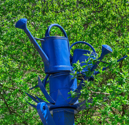 Blue watering cans made of metal stacked on a blue post amidst a green bush.の写真素材