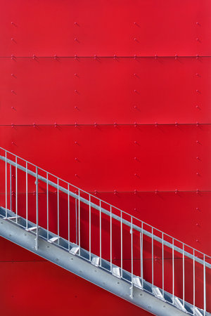 Metal staircase outdoors in front of a red wall, clad with large square panels