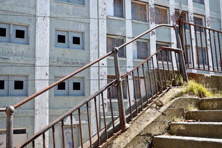 A Crumbling Stairway with Rusty Railing:  An image in an urban setting highlighting damaged steps and rusted iron railing in the foreground.  In the background is the facade of an old concrete building showing decay with boarded windows.の写真素材