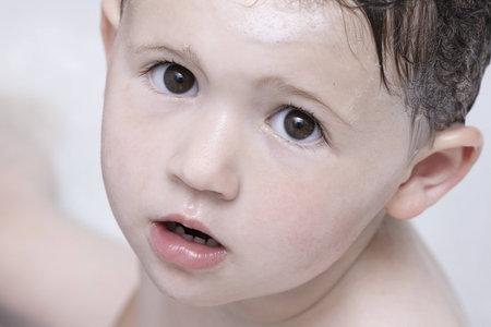 A toddler in the bathtub with big brown eyes looking directly into the camera   His has wet hair and there are bubbles on his head   The image is a tight crop to the face の写真素材