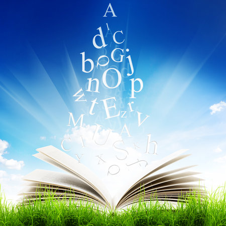 Open book with flying letters in green grass over blue sky background. Magic bookの写真素材