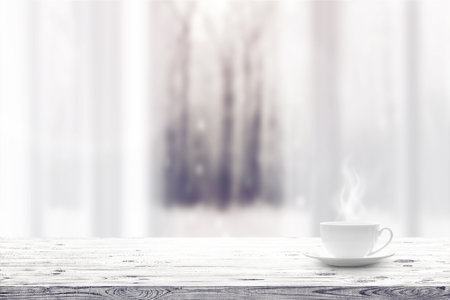 Cup with hot drink on wooden table and window over winter snow covered forest backgroundの写真素材
