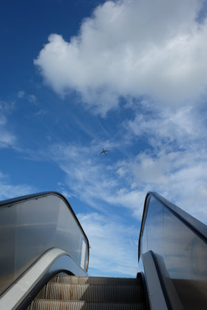 Open air escalator raise towards sunny blue sky with white clouds. An airplane flying in the sky. freedom, success, promotion conceptの写真素材
