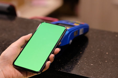 one hand using green screen phone close to POS terminal to pay on counter. Mobile payment