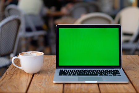 close up one green screen laptop computer on wooden table with a cup of coffee. blur sidewalk cafeの写真素材
