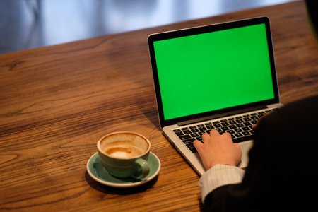 over the shoulder view of woman using green screen laptop computer with coffee in cafe.