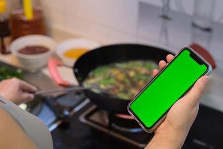 over the shoulder of woman hand holding green screen phone when cooking food in kitchenの写真素材