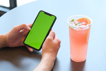 close up hand holding green screen smart phone with a cup of cold fruit drink on white table