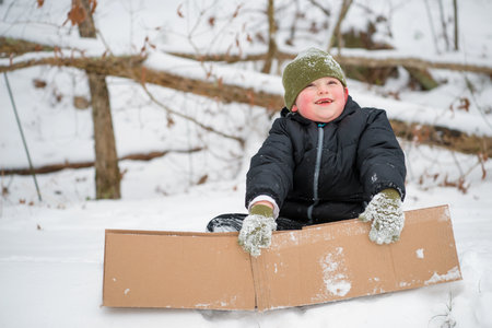 Child playing in snow using cardboard box to slide down hill