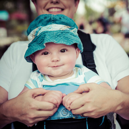 Baby boy riding in infant carrier by mother outdoors at crowded park in vintage filtered imageの写真素材