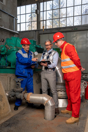 Technical Manager with Digital Tablet Talking to Factory Workers in Mechanical Room. District Heating Power Plant. Digital Technology and Teamwork Concept. Industry 4.0の写真素材