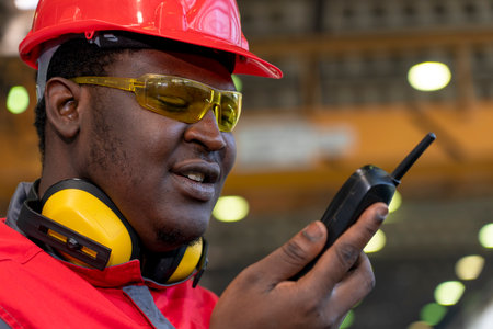 Black Worker In Protective Workwear Talking On Radio Communication Equipment In A Factory. Portrait...
