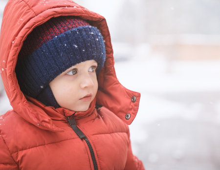 Baby boy while it snows looks towards the emptiness, covered with red winter jacket and woolen hat, close-up.の写真素材