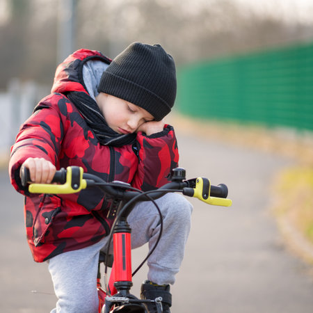 Sad, angry boy with hat and scarf sitting on bicycle at park.の写真素材