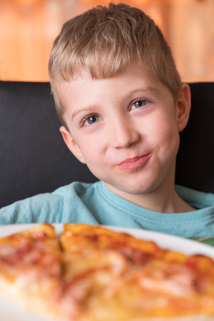 Beautiful happy young boy smiling and biting off slice of fresh made pizza, he sit at black chair, He has blonde hair.の写真素材