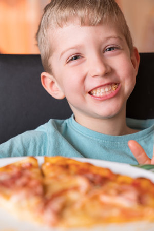 Beautiful happy young boy smiling and biting off slice of fresh made pizza, he sit at black chair, He has blonde hair.の写真素材