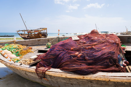 Wooden fisherman boats on sandy beach, in the foreground red fishing nets, background dhow boat and sea, Zanzibar, Tanzaniaの写真素材