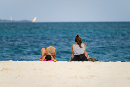 Two young women relax on the sandy beach, one reads a book, blue ocean backgroundの写真素材