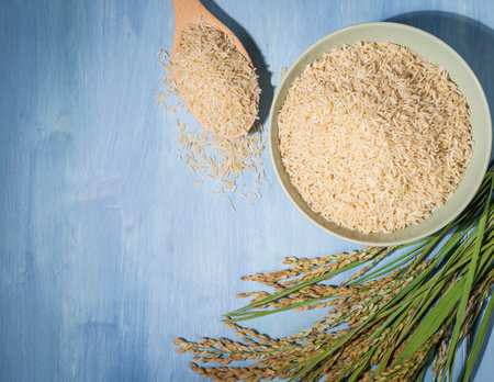 A bowl of brown rice is placed on a blue table next to a spoonful of rice and ears of rice.The bowl...