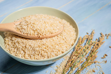 A bowl of brown rice is placed on a blue table next to a spoonful and ears of rice.The raw rice is spread out in the bowl and the spoon is full of raw-rice and sitting on top of itの写真素材