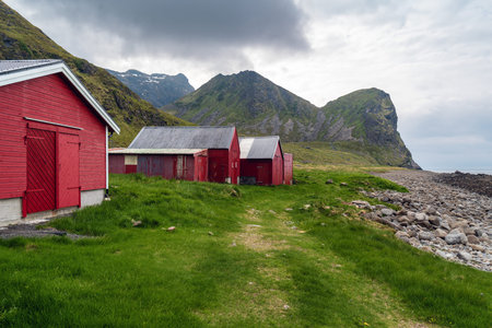 Red wooden cabins standing on a green coastal meadow with rocky shoreline and dramatic mountain landscape at Unstad Beach on the Lofoten Islands, Norway. Nordic coastal scenery under overcast sky.の写真素材